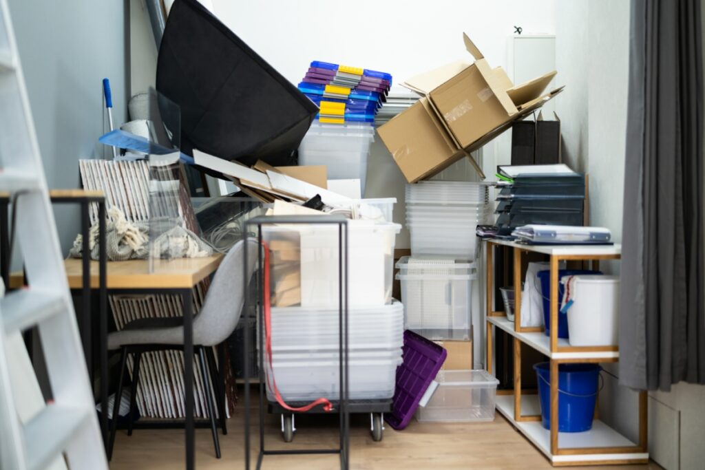 A cluttered room filled with stacked plastic containers, cardboard boxes, a table with chairs, various office supplies, and a black foldable chair leaning on a pile of cardboard boxes—awaiting the expertise of hauling services for a much-needed clear-out.